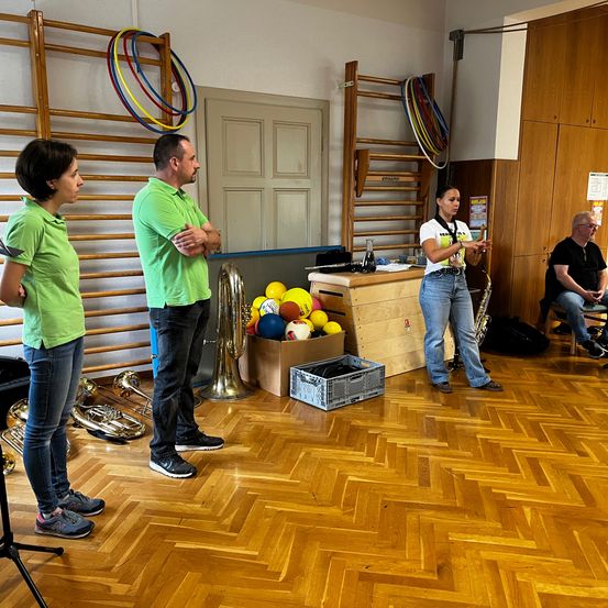 Drei Personen in grünen Shirts stehen in einem Raum mit Holzböden und Musikinstrumenten. Eine Frau spielt Saxophon, während ein Mann und eine Frau in der Nähe stehen.
