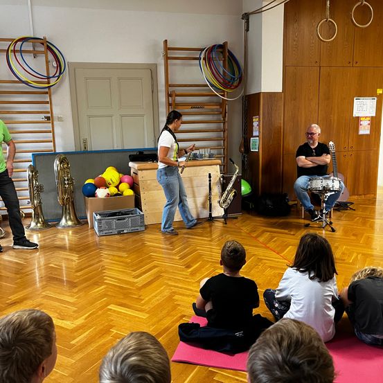 Eine Gruppe von Kindern sitzt auf einer Yogamatte und beobachtet eine Frau, die Flöte spielt. Zwei Männer spielen Saxophon und Schlagzeug. Verschiedene Instrumente sind im Raum ausgestellt.