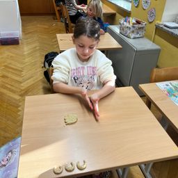 Ein junges Mädchen sitzt in einem Klassenzimmer an einem Schreibtisch und arbeitet mit Keksen, während zwei andere Kinder in der Nähe sind.