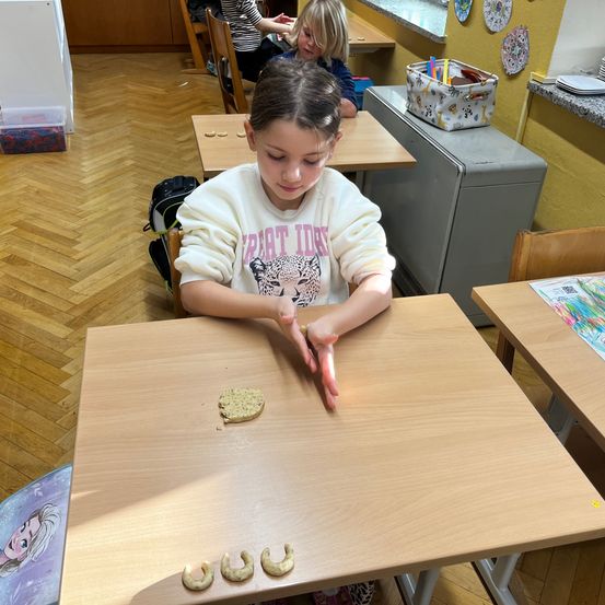 Ein junges Mädchen sitzt in einem Klassenzimmer an einem Schreibtisch und arbeitet mit Keksen, während zwei andere Kinder in der Nähe sind.