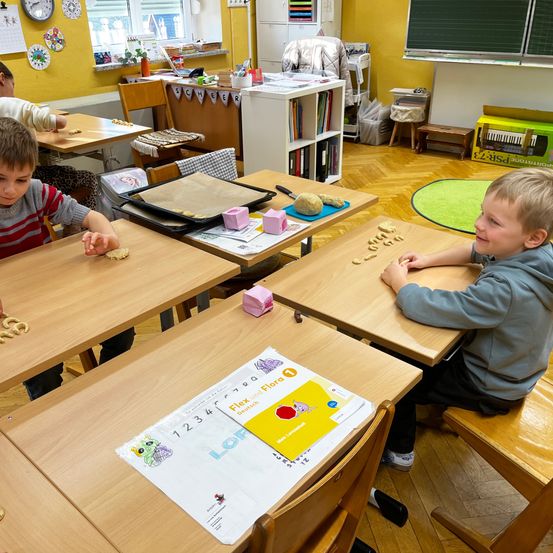 Zwei Jungen sitzen an Schreibtischen in einem Klassenzimmer und sind in eine Aktivität mit Lebensmitteln verwickelt. Ein Junge lächelt und der andere schaut auf sein Essen. Die Schreibtische haben Tabletts mit Lebensmitteln und der Raum hat gelbe Wände und Holzböden.
