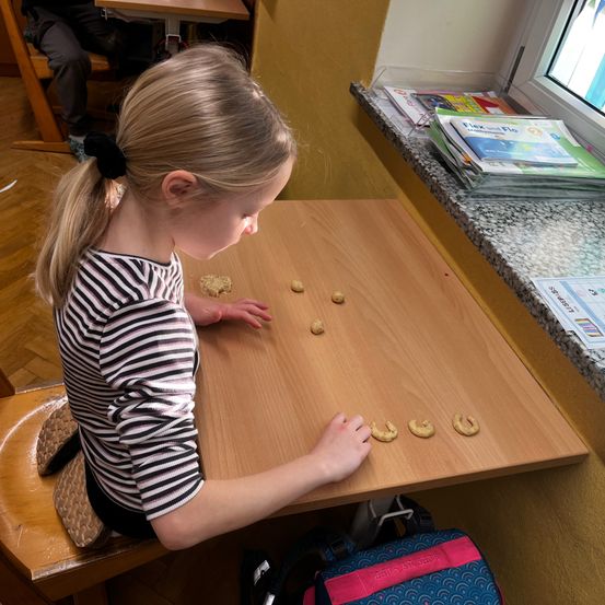 Ein kleines Mädchen sitzt an einem Holztisch und ordnet Kekse in einer Reihe an. Sie trägt ein gestreiftes Shirt. Ein Rucksack und Bücher sind in der Nähe.