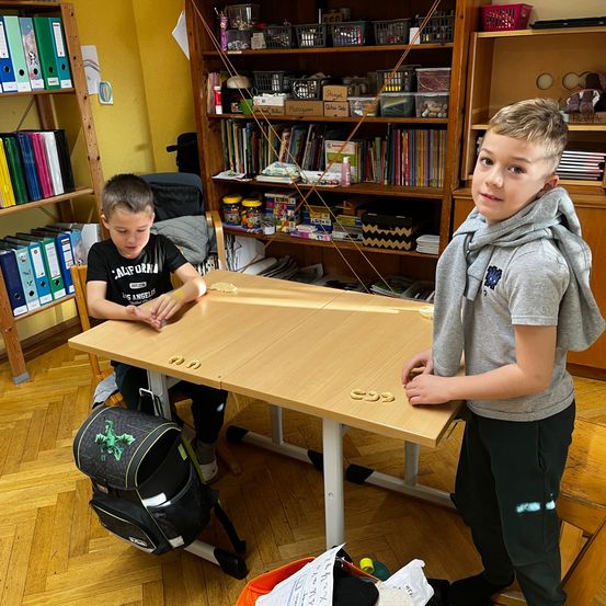 Zwei Jungen spielen ein Brettspiel auf einem Holztisch. Ein Junge sitzt und der andere steht neben ihm. Hinter ihnen befinden sich Regale voller Bücher und Spielzeug.