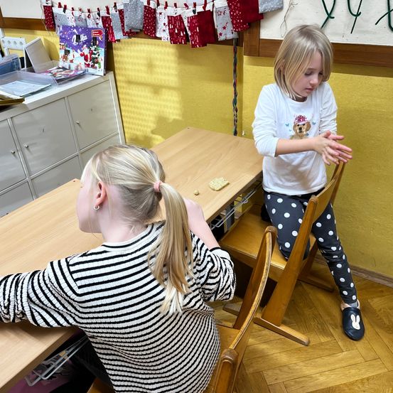 Zwei junge Mädchen in einem Klassenzimmer, eine sitzt an einem Schreibtisch, während die andere in der Nähe steht und möglicherweise ein Spiel spielt. Auf dem Schreibtisch befindet sich Essen.