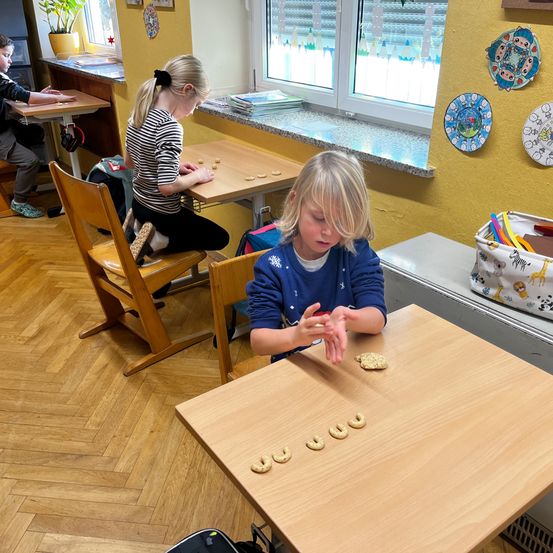 Zwei Mädchen sitzen in einem Klassenzimmer, eine konzentriert sich auf ihr Bastelprojekt, während die andere mit Keksen am Tisch spielt. Ein Junge sitzt in der Ecke.
