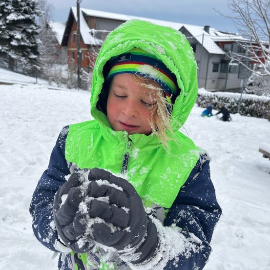 Ein Kind in einem grünen und blauen Mantel spielt im Schnee. Andere Kinder spielen im Hintergrund.