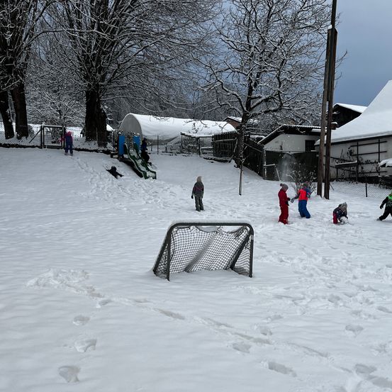 Mehrere Kinder spielen im Schnee, einige rutschen einen schneebedeckten Hang hinunter und andere spielen mit einem Hockeynetz.