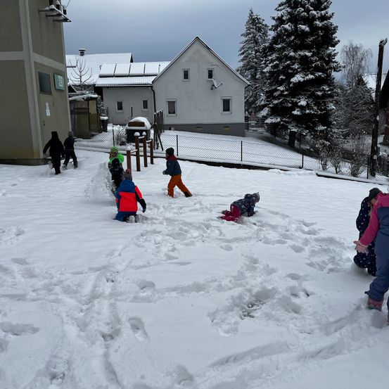 Eine Gruppe von Kindern spielt im Schnee vor einem Gebäude. Sie tragen Winterkleidung und einige bauen Schneemänner.