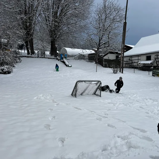 Kinder spielen im Schnee auf einem Hügel mit einer Rutsche und einem Tor. Ein Haus mit schneebedecktem Dach ist im Hintergrund.