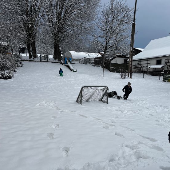 Kinder spielen im Schnee auf einem Hügel mit einer Rutsche und einem Tor. Ein Haus mit schneebedecktem Dach ist im Hintergrund.