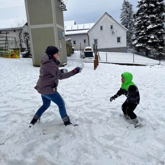 Eine Frau und ein Kind spielen im Schnee und tragen Winterkleidung. Ein Haus mit einem Zaun und ein schneebedeckter Baum sind im Hintergrund zu sehen.