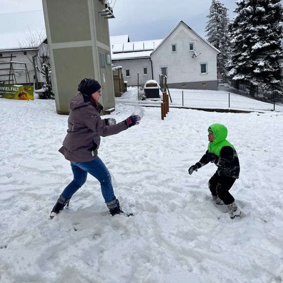 Eine Frau und ein Kind spielen im Schnee und tragen Winterkleidung. Ein Haus mit einem Zaun und ein schneebedeckter Baum sind im Hintergrund zu sehen.