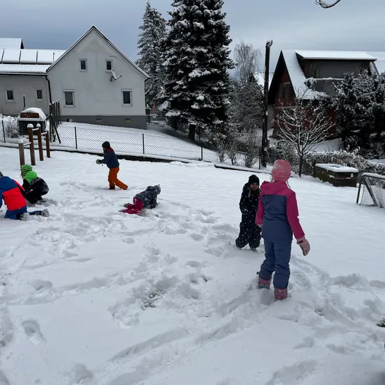 Eine Gruppe von Kindern spielt im Schnee vor einem Haus mit Zaun und verschneiten Bäumen.