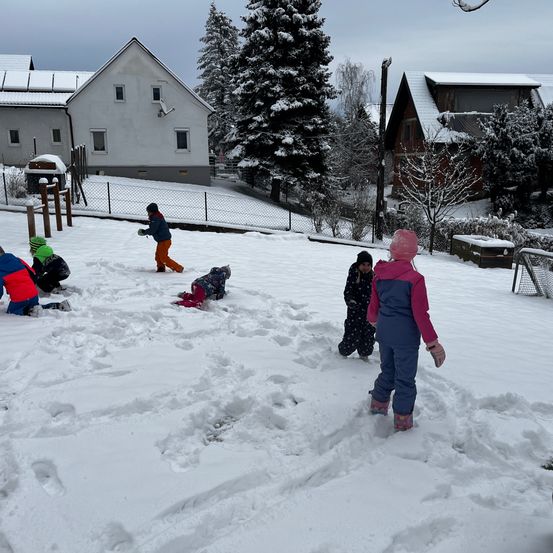 Eine Gruppe von Kindern spielt im Schnee vor einem Haus mit Zaun und verschneiten Bäumen.