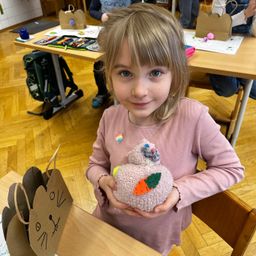 Ein kleines Mädchen in einem rosa Langarmshirt hält ein Kuscheltier in einem Klassenzimmer. Ein Beutel mit Katzendesign steht auf dem Tisch vor ihr.