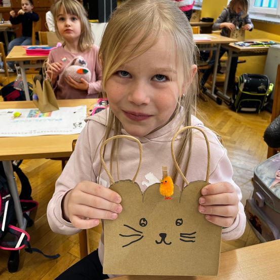 Ein kleines Mädchen im Klassenzimmer, das einen Papierbeutel in Katzenform mit einem gelben Spielzeug oben hält. Hinter ihr hält eine andere Mädchen ein Plüschtier, und andere sitzen an Schreibtischen.