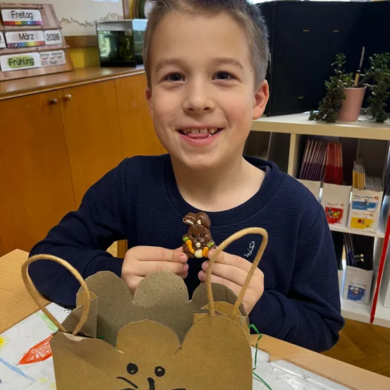 Ein Junge hält einen Schokoladenosterhasen in einer Papptüte. Er sitzt an einem Tisch in einem Klassenzimmer mit Regalen voller Bücher und Pflanzen dahinter.