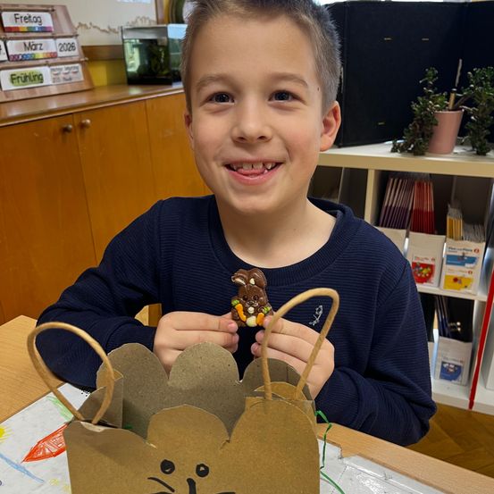 Ein Junge hält einen Schokoladenosterhasen in einer Papptüte. Er sitzt an einem Tisch in einem Klassenzimmer mit Regalen voller Bücher und Pflanzen dahinter.