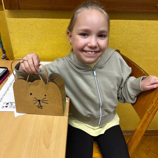 Ein junges Mädchen in einer grauen Kapuzenjacke hält einen braunen Papierbeutel mit einem Katzengesicht darauf. Sie sitzt an einem Tisch mit einer gelben Wand im Hintergrund.