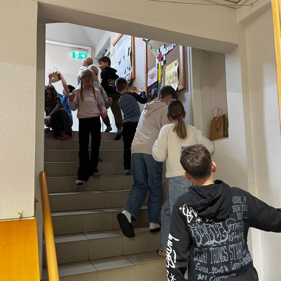 Eine Gruppe von Kindern, die Papiertüten halten, klettert die Treppe hinauf. Ein Kind in einem rosa Shirt schaut zur Kamera. Ein Junge mit Kapuzenpullover ist unten. An der Wand sind Papiere befestigt.