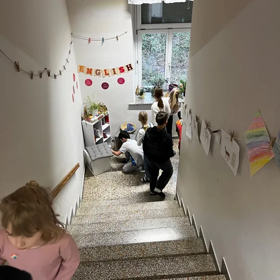 Kinder stehen auf den Treppen, manche stehend, andere sitzend oder kniend. Ein weißer Regal mit Topfpflanzen befindet sich links. Ein Banner mit dem Wort Englisch hängt darüber.