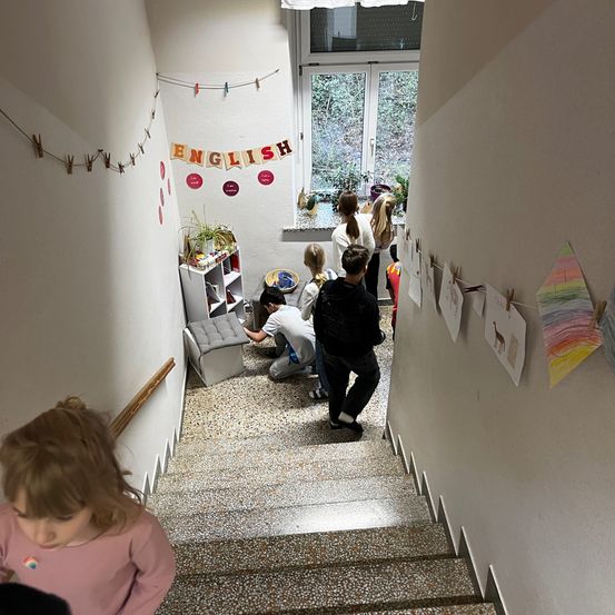 Kinder stehen auf den Treppen, manche stehend, andere sitzend oder kniend. Ein weißer Regal mit Topfpflanzen befindet sich links. Ein Banner mit dem Wort Englisch hängt darüber.