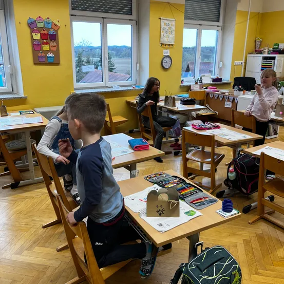In einem gelb gestrichenen Klassenzimmer sitzen Kinder an Tischen mit Kunstmaterialien. Ein Junge sitzt vorne, während ein Mädchen in der Nähe einen Becher hält. Der Raum verfügt über Holzböden, gelbe Wände und Fenster mit einer malerischen Aussicht.