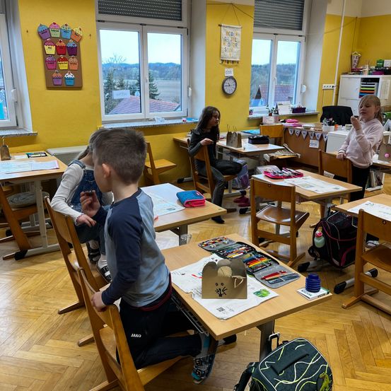 In einem gelb gestrichenen Klassenzimmer sitzen Kinder an Tischen mit Kunstmaterialien. Ein Junge sitzt vorne, während ein Mädchen in der Nähe einen Becher hält. Der Raum verfügt über Holzböden, gelbe Wände und Fenster mit einer malerischen Aussicht.