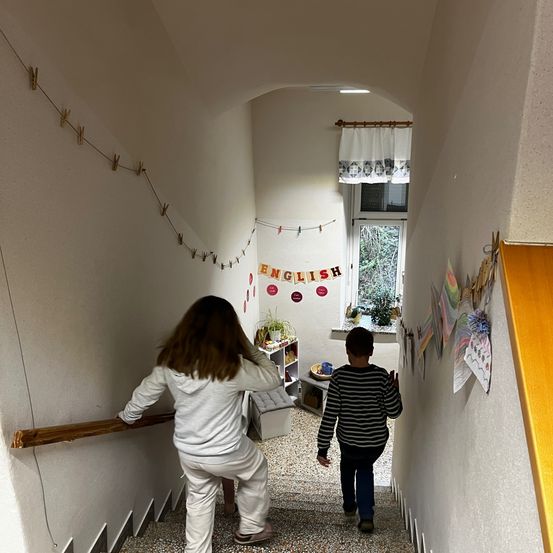 Zwei Kinder gehen eine Treppe hinauf. Ein Mädchen in Weiß hält das Geländer, während ein Junge im gestreiften Hemd hinter ihr geht. Der Flur ist mit bunten Papieren dekoriert.
