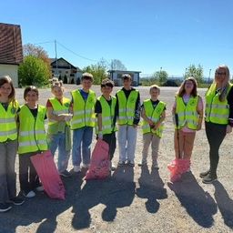 Eine Gruppe von Kindern in gelben Westen, mit rosa Tüten in der Hand, steht auf Kies mit Bäumen und Gebäuden im Hintergrund.