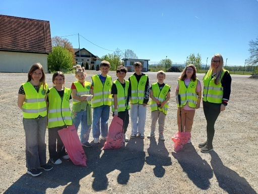 Eine Gruppe von Kindern in gelben Westen, mit rosa Tüten in der Hand, steht auf Kies mit Bäumen und Gebäuden im Hintergrund.