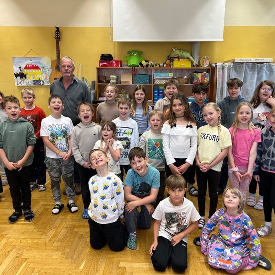 Gruppenfoto von Kindern und einem erwachsenen Mann, die in einem Klassenzimmer lächeln. Im Hintergrund befinden sich verschiedene Spielzeuge und eine Gitarre.