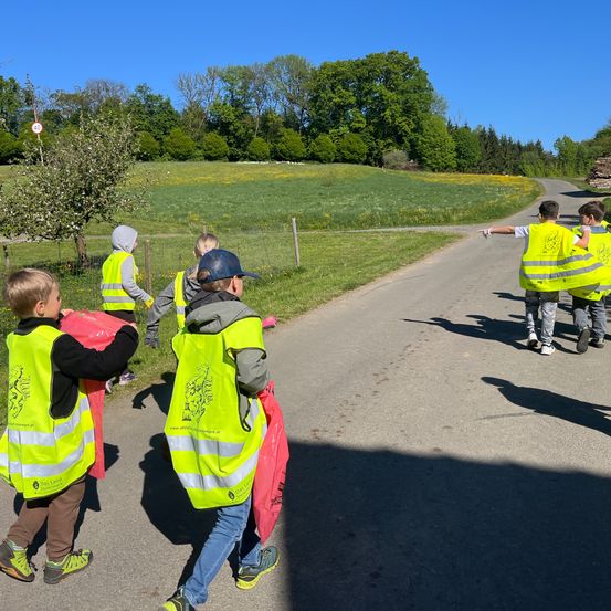Eine Gruppe von Kindern in neonfarbenen Westen und mit roten Taschen läuft auf einem Landweg. Im Hintergrund befinden sich Bäume und ein Feld.