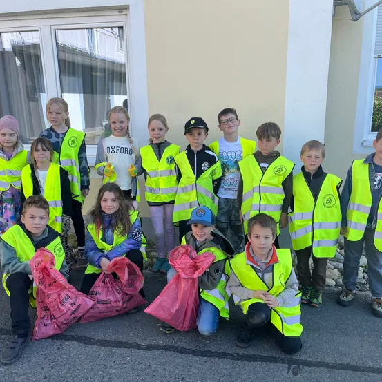 Eine Gruppe von Kindern, die neongrüne Sicherheitswesten tragen und Plastiktüten halten, posiert für ein Foto vor einem Gebäude.