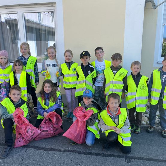 Eine Gruppe von Kindern, die neongrüne Sicherheitswesten tragen und Plastiktüten halten, posiert für ein Foto vor einem Gebäude.