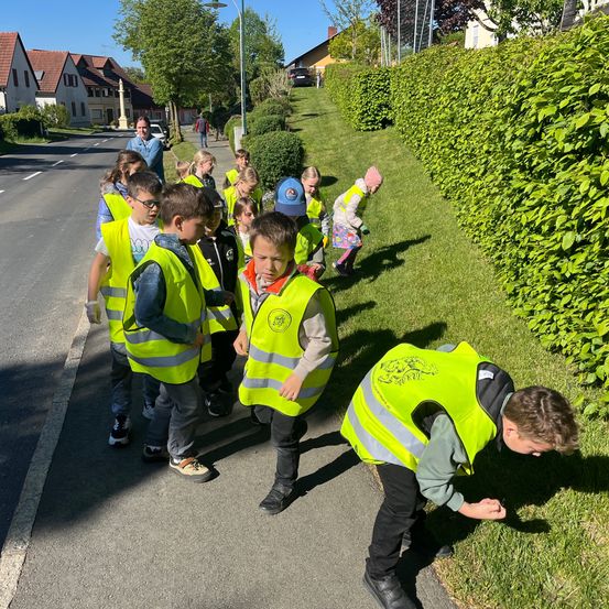Eine Gruppe von Kindern in Warnwesten geht am Straßenrand entlang. Ein Kind beugt sich herunter, um etwas am Boden zu untersuchen.