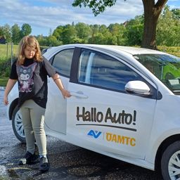 Ein junges Mädchen steht neben einem weißen Auto mit einem Schild, das Hallo Auto! steht. Das Auto ist auf einer Straße geparkt, mit einem Baum und einem Zaun im Hintergrund.
