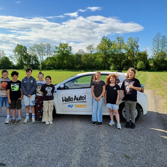 Eine Gruppe von Kindern und einer Frau posiert neben einem weißen Auto mit der Aufschrift Hallo Auto!. Sie stehen auf einem Schotterweg mit einem Grasfeld und Bäumen hinter ihnen.