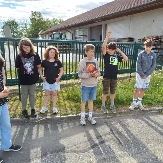 Fünf junge Personen stehen neben einem grünen Zaun. Einer hält einen Basketball, ein anderer gestikuliert mit einer Hand. Sie tragen Freizeitkleidung, eine Person trägt eine Brille. Hinter ihnen steht ein Gebäude mit einem braunen Dach und einem großen offenen Bereich mit einem Traktor.