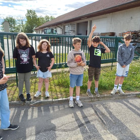 Fünf junge Personen stehen neben einem grünen Zaun. Einer hält einen Basketball, ein anderer gestikuliert mit einer Hand. Sie tragen Freizeitkleidung, eine Person trägt eine Brille. Hinter ihnen steht ein Gebäude mit einem braunen Dach und einem großen offenen Bereich mit einem Traktor.