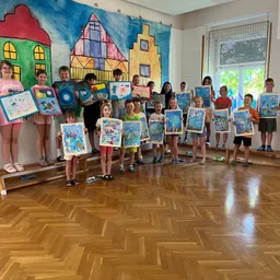 Eine Gruppe von Kindern steht auf einer Bühne in einem Raum, hält verschiedene farbenfrohe Gemälde in den Händen, mit einem Wandgemälde hinter ihnen.