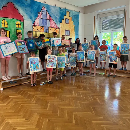 Eine Gruppe von Kindern steht auf einer Bühne in einem Raum, hält verschiedene farbenfrohe Gemälde in den Händen, mit einem Wandgemälde hinter ihnen.