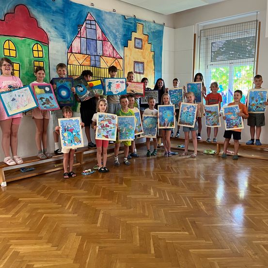 Eine Gruppe von Kindern steht auf einer Bühne in einem Raum, hält verschiedene farbenfrohe Gemälde in den Händen, mit einem Wandgemälde hinter ihnen.