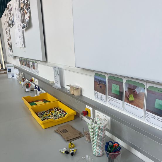 Ein Klassenzimmer-Setup mit einem Schreibtisch und Regalen mit Bastelzubehör und Bildern an der Wand. Ein gelber Behälter mit Perlen und ein durchsichtiger Behälter mit Strohhalmen stehen auf dem Tisch.