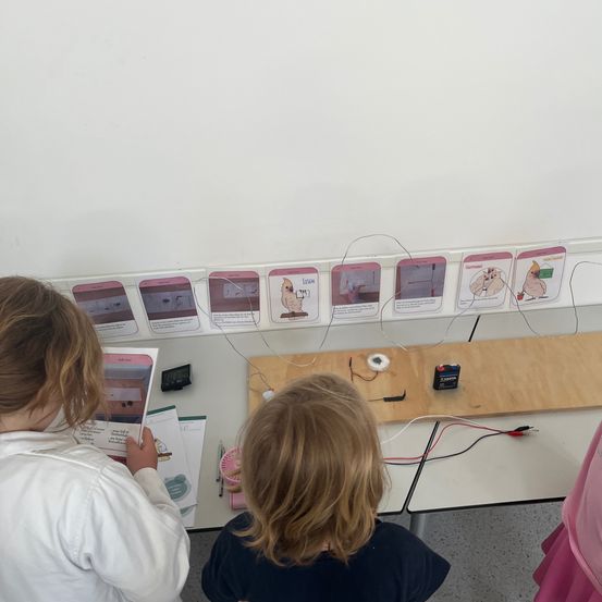 Zwei junge Kinder sind in einem Klassenzimmer und arbeiten an einem Wissenschaftsprojekt mit einem elektrischen Kreislauf. Eine Tafel ist im Hintergrund.