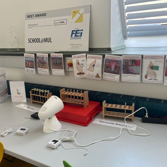 Ein Laboraufbau mit einem weißen Tisch, Kabeln und einem kleinen weißen Gerät. Ein Poster mit dem Text IMST AWARD ist an der Wand.
