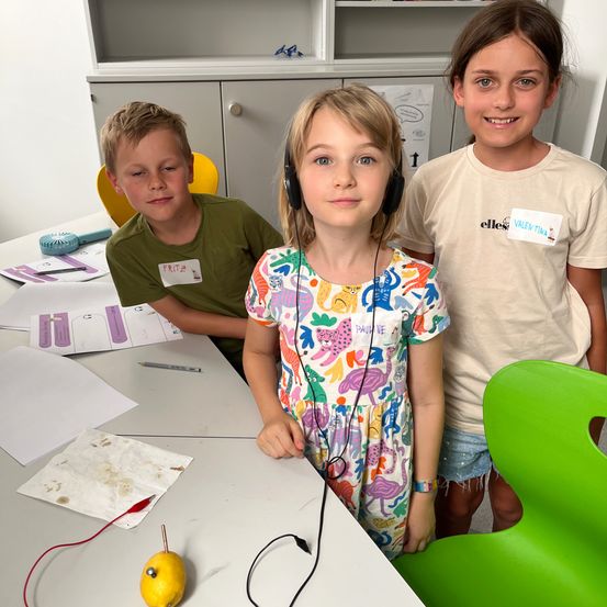 Drei Kinder in einem Klassenzimmer. Ein Mädchen trägt Kopfhörer und steht in der Mitte. Zwei andere Kinder sitzen hinter ihr. Ein Schreibtisch mit Papieren, einem Bleistift und einer Zitrone steht vor ihnen.