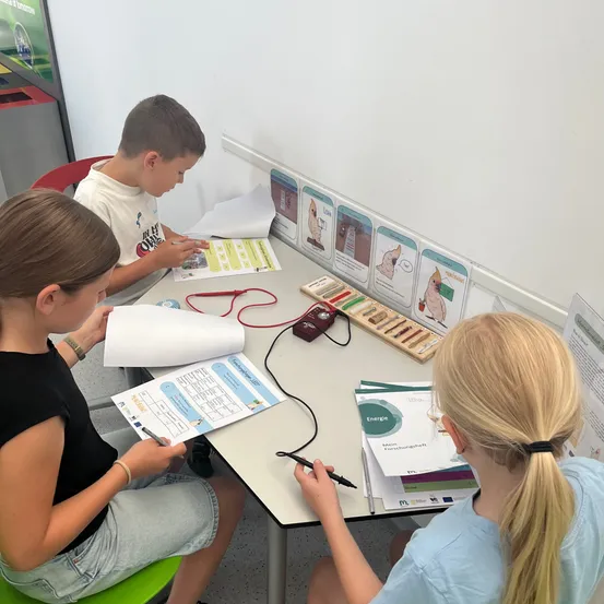 Drei Kinder sitzen an einem Tisch. Ein Junge konzentriert sich auf ein Papier mit einem roten Draht, während zwei Mädchen auf Papieren schreiben. Hinter ihnen befinden sich Poster an der Wand.