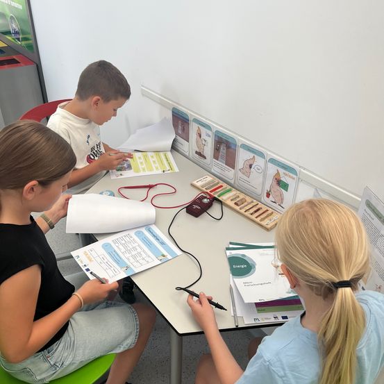 Drei Kinder sitzen an einem Tisch. Ein Junge konzentriert sich auf ein Papier mit einem roten Draht, während zwei Mädchen auf Papieren schreiben. Hinter ihnen befinden sich Poster an der Wand.