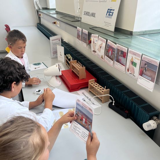 Drei Kinder sind in einem Klassenzimmer. Sie machen ein wissenschaftliches Experiment mit einer Lampe, einer roten Box und einer Holzstruktur. Sie tragen weiße Mäntel und halten eine Karte.